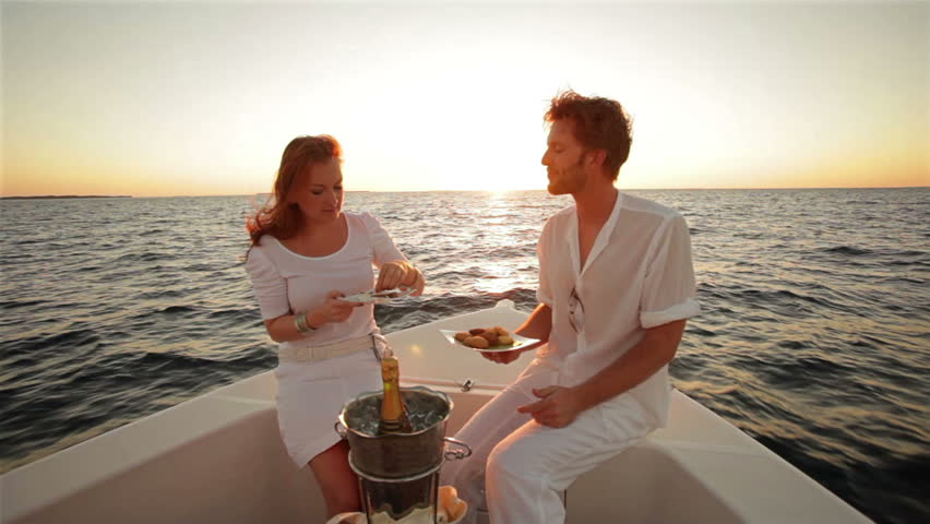 young couple in white sitting on boat at sunset with champagne and appetizer 