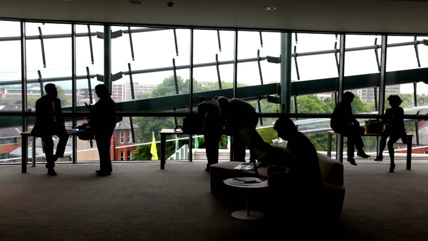 People standing near the windows, one man is sitting on the sofa near the table.