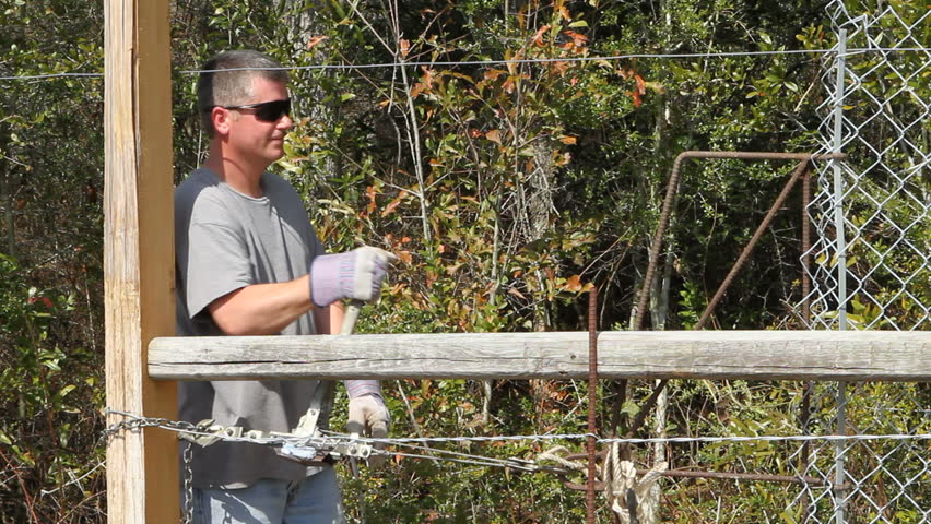 Laborer uses a come-along winch to stretch a length of chain link fence into position.