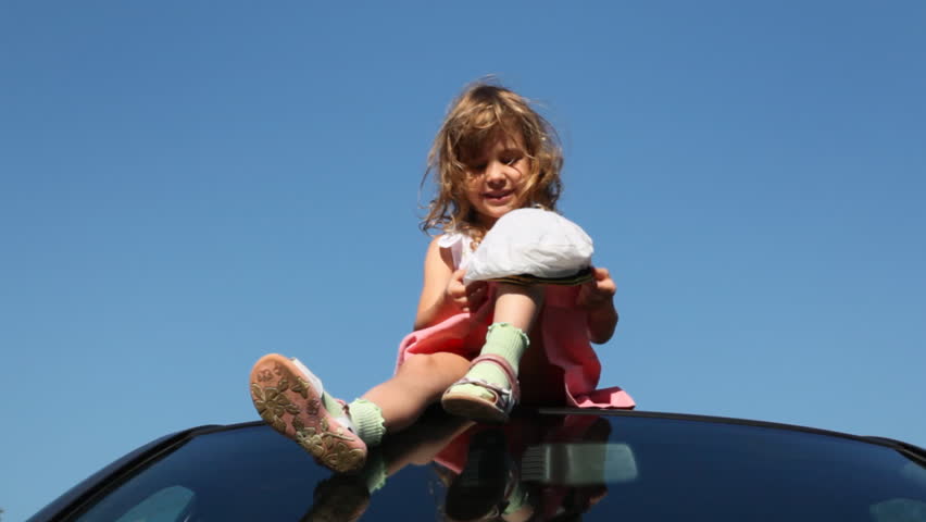 nice little girl sits at roof of car against blue sky, puts big captain's cap and turns imaginary driving wheel