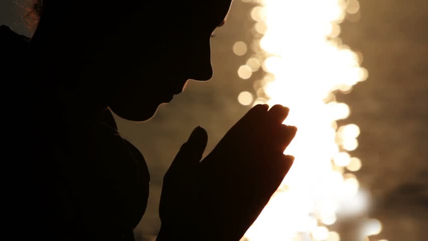 Silhouette of woman head with sunshine reflected in water behind, holding hand in namaste
