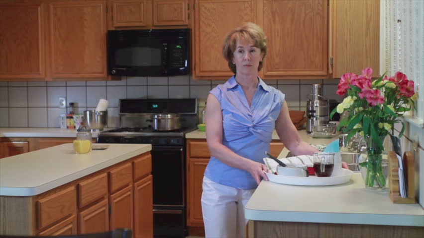 A woman carries a tray with soup, tea, and other items on it out from the kitchen and then walks off camera.