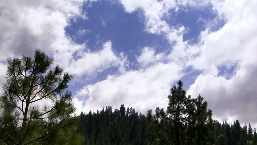 Time lapse clouds sail across a pale blue sky over pine-studded hills.