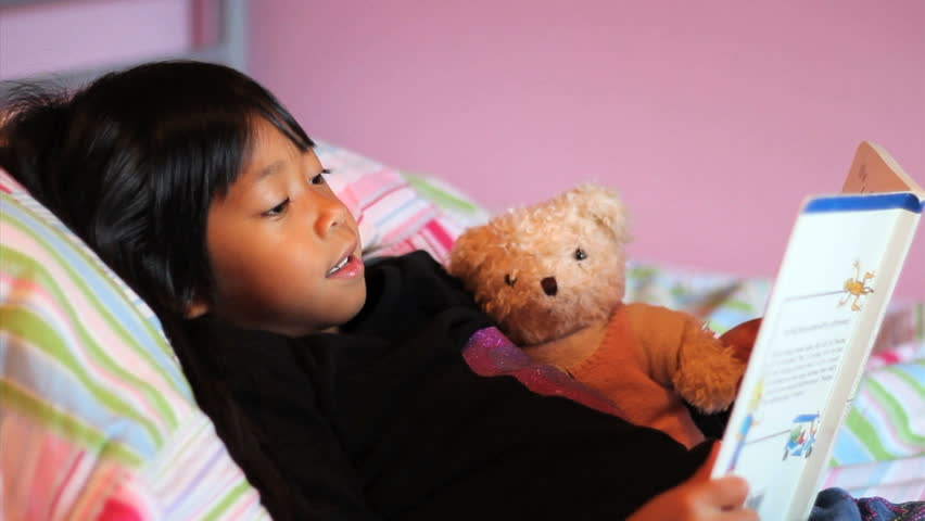 A cute little 5 year old Asian girl reads a story book to her teddy bear.