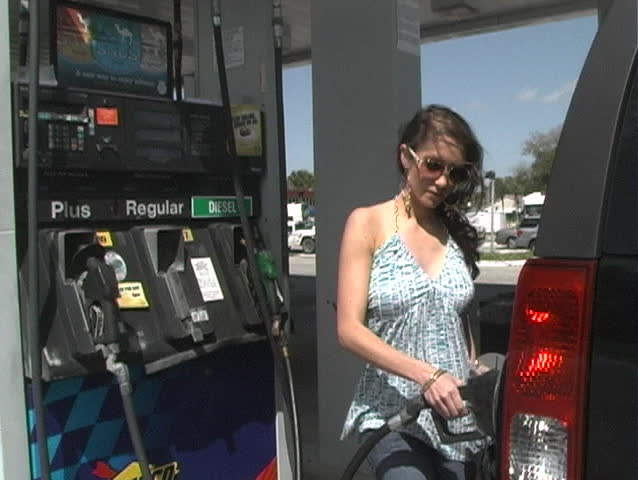 A beautiful brunette finishes gassing up her vehicle, replaces the gas pump nozzle, replaces her gas cap, and walks out of view.  See additional clips from alternate angles to form a series.