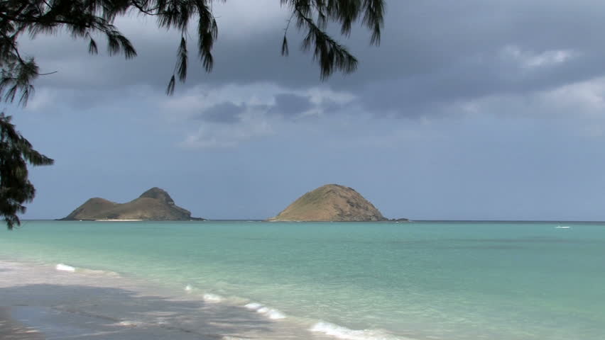 Tropical beach on Oahu, Hawaii. Islands off shore in Pacific ocean. Outrigger paddling by distance islands. Waves on white sand with tree limbs overhead. Scenic, relaxing, calm seascape.