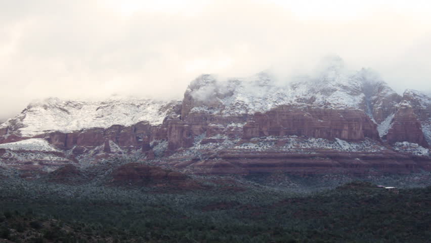 Sunlight shines down through heavy, early-morning clouds covering the mountains of Sedona, Arizona with a light dusting of snow. Wide shot.
