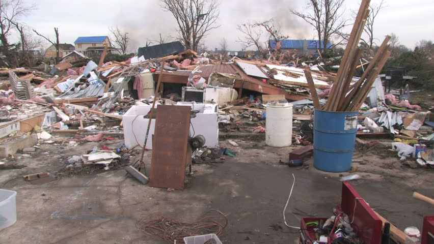 Looking over the chaotic wreckage of a demolished house after a tornado, with smoke rising in the background. 