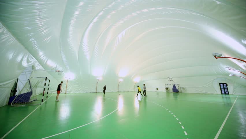 Four students play soccer on football field under cupola in inflatable gymnasium, view from corner