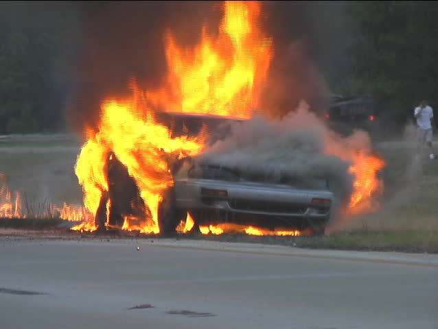 An abandoned import car on fire burns and later has something small explode while the horn blares.