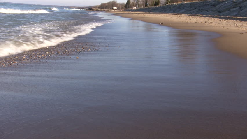 Waves gently hit the beach, then recede as the sunlight illuminates the surf and the blue sky reflects off of the wet sand. (High Definition)
