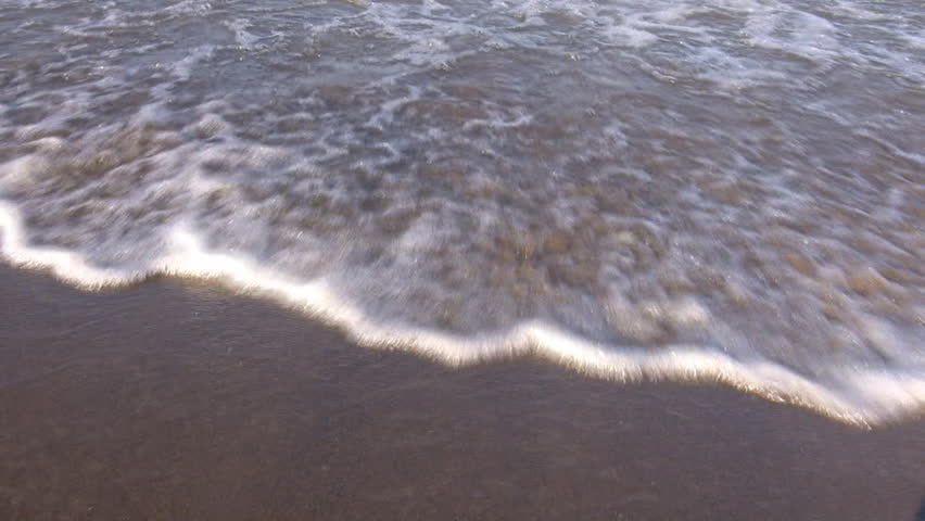 Waves gently hit the beach towards and then under the frame, then gently recede as the sunlight reflects and glitters off of the water and sand. (High Definition)