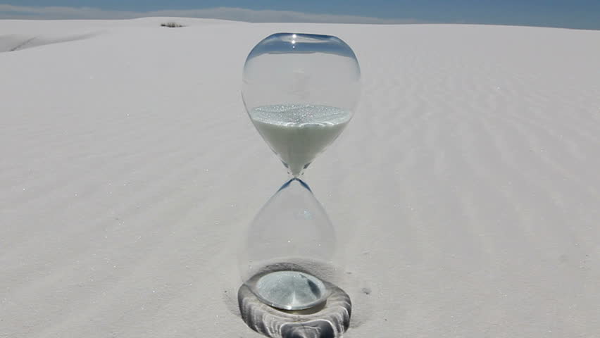 Time Lapse, White sand spills through hourglass positioned on white, textured sand dunes, blue sky. 1080p