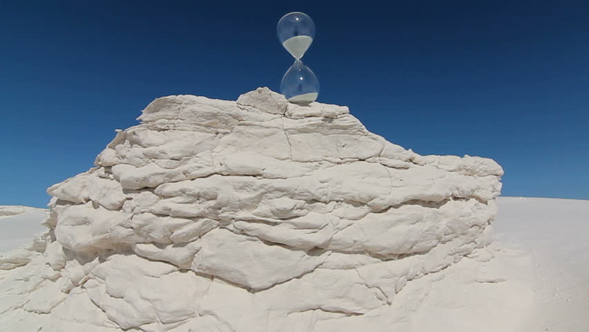 Sand seeps through hourglass perched on large, textured mound of white sand rising from landscape, blue sky background. 1080p
