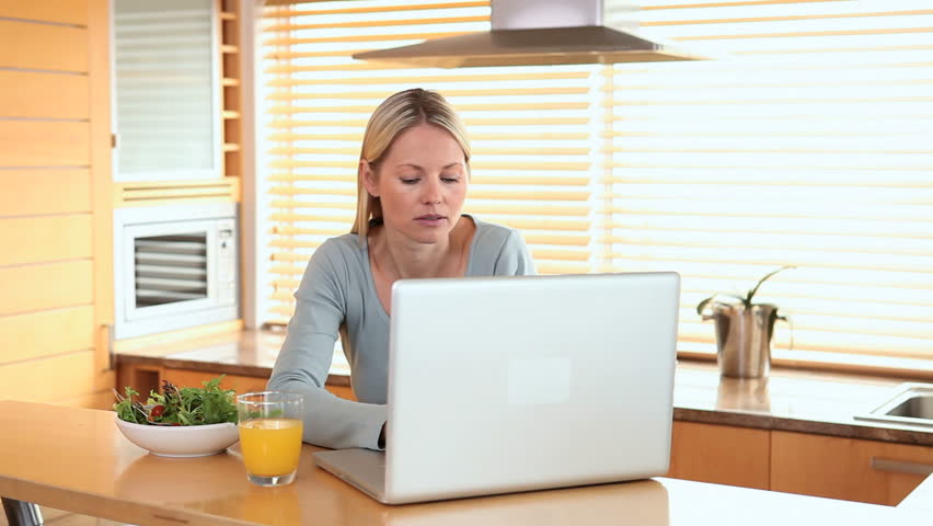 Woman drinks a glass of orange juice while looking at a laptop screen
