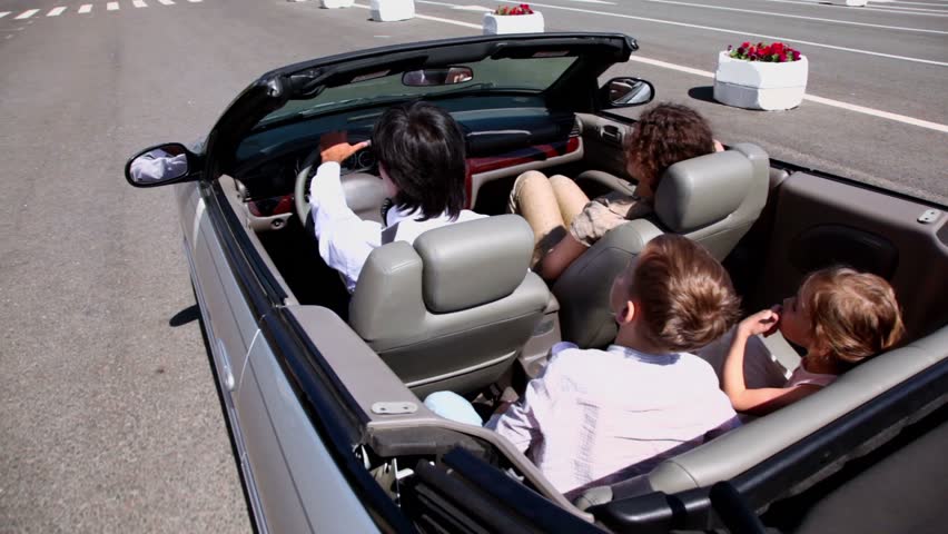 Parents and two kids sit in cabriolet and ride by road at sunny summer day