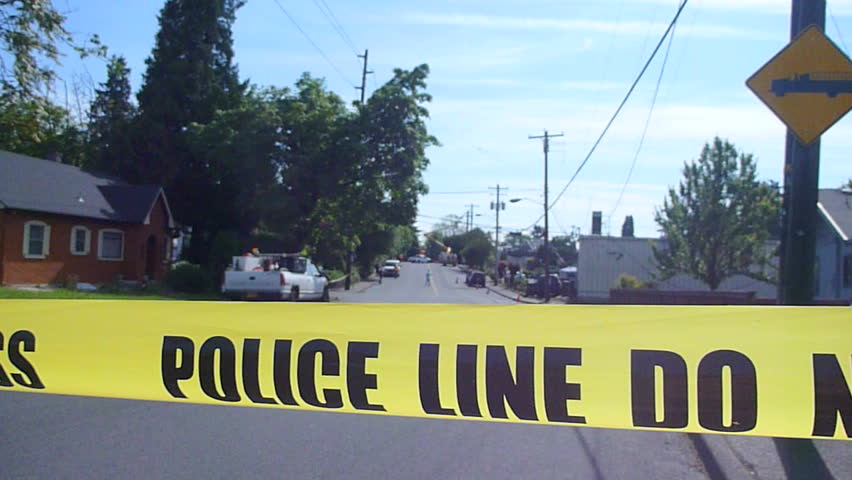 Police caution line reads Do Not Cross in residential neighborhood street blocking traffic at crime scene.