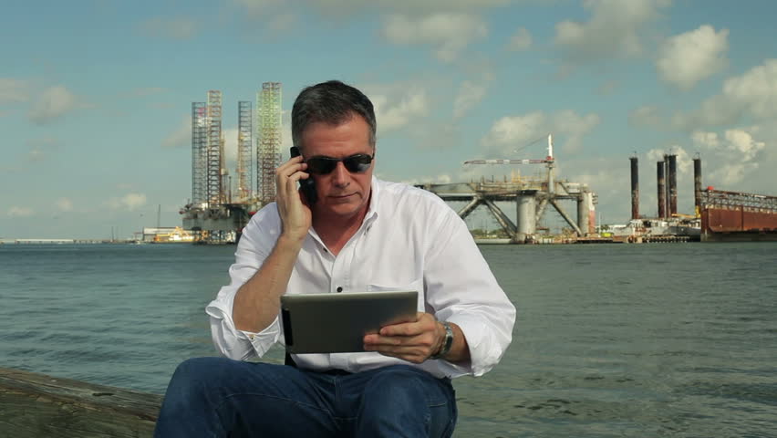 A man sitting on a pier with drilling platforms in the background using a cell phone and electronic tablet to conduct business.
