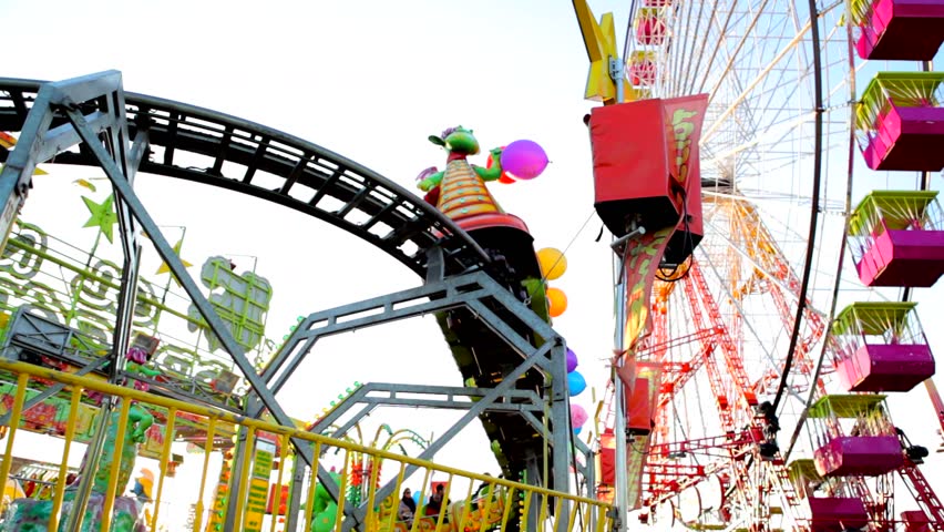 Colorful ferris wheel and fast train rides in motion at a theme park against a blue sky.