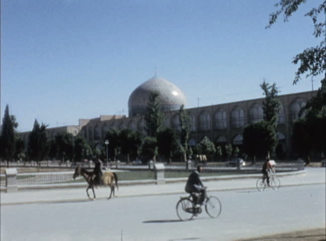 ISFAHAN, IRAN - JULY 1956: Men riding horses and bikes in Naghshi-i Jahan Square with 17th century Masjid-i-Shah Islamic Royal Blue Mosque in background in Isfahan, Iran. Shot on 16mm film.