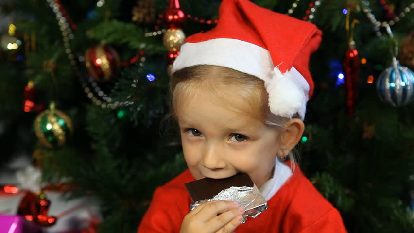 Child Eating Chocolate Near Christmas Tree, Little Girl Dressed in Santa Claus Eating Chocolate by the Christmas Tree, Close Up