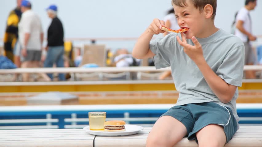 boy sits and eats pizza, washing down with water against people of vacationers on chaise lounges