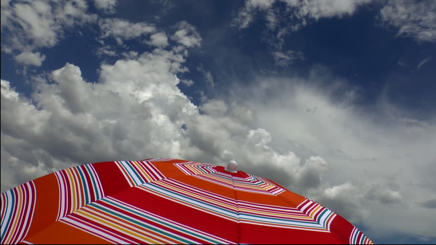 Time Lapse, Thick, fluffy clouds soar above colorful, striped umbrella in sunny, bright, blue sky. 1080p