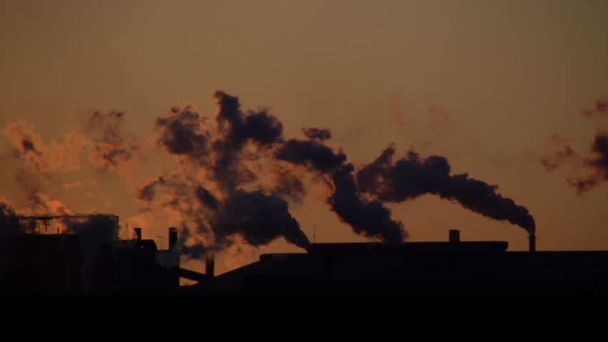 The exhaust from a factory is backlit by the sun during sunset. (High Definition)