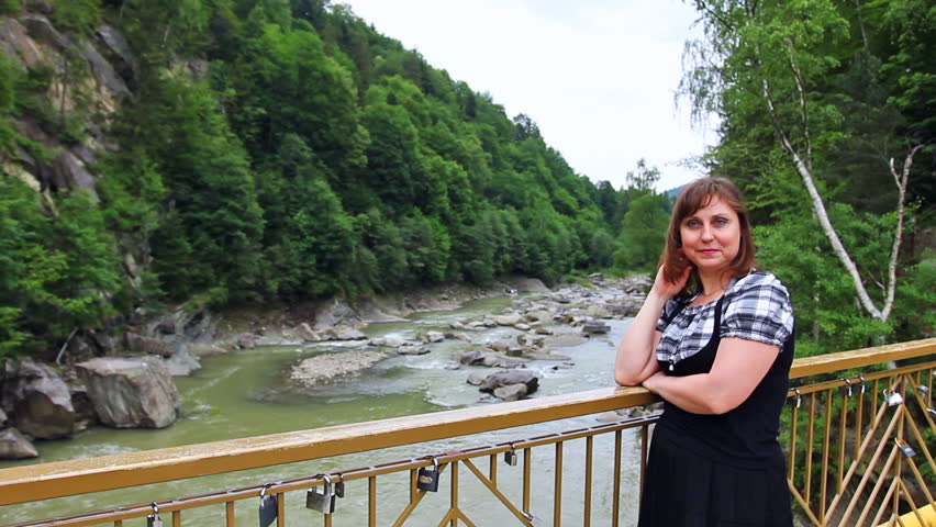 pretty woman stands near railing bridge, then looks on river in Carpathian Mountains