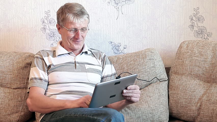 Senior adult Caucasian man interested with a new tablet pc, sitting on sofa in domestic room