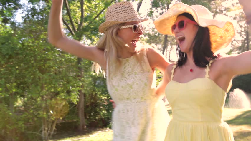 Two women wearing sunglasses and straw hats dance along country path.