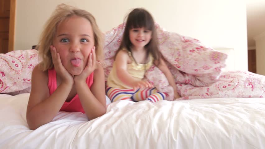 Two girls hiding under sheet in bed before emerging and pulling funny faces.