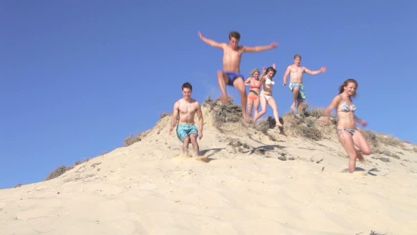 Group of teenage friends appear over peak ofdune before running down and jumping past camera.