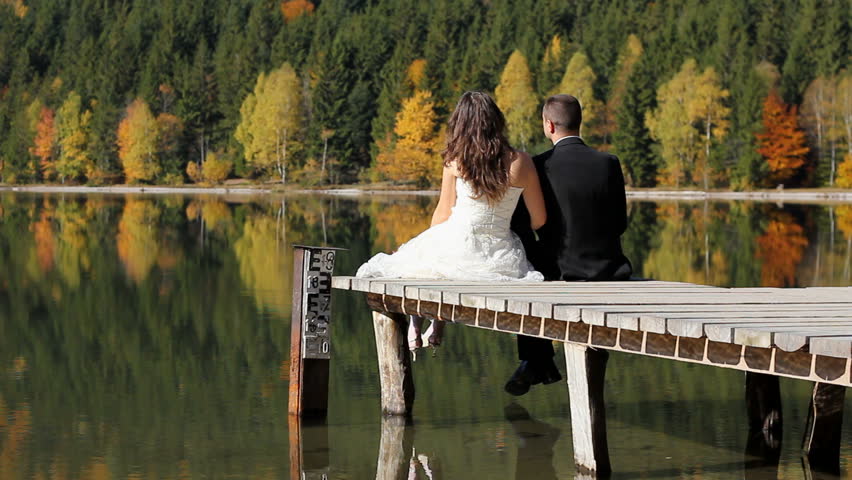 Just married couple with a life in front of them, bride still wearing wedding dress and groom in black suit, sitting together on a wooden pontoon and admiring the superb nature autumn scenery