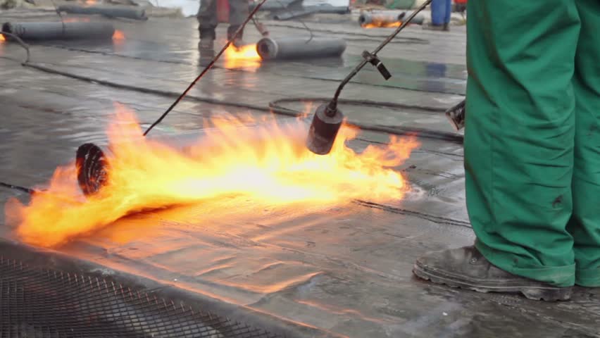 Labourers use burners to warm ruberoid for waterproofing and roll up it by hooks, closeup view