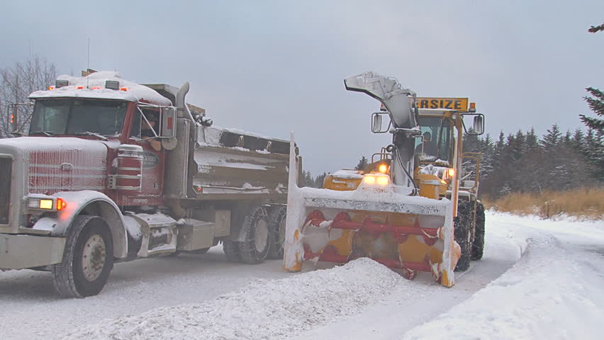 HOMER, AK CIRCA 2012: Snow blower on an excavator paired with a dump truck to clean plow piles on snowy city street.