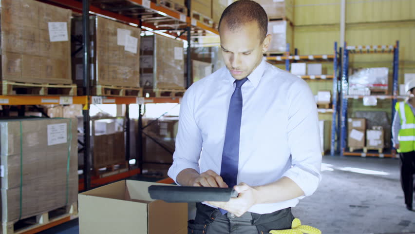 A young man is working in a warehouse with a digital tablet, he is dressed in a shirt and tie. He gives instructions to a female worker and another employee can be seen working in the background