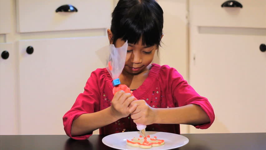 A cute little 6 year old Asian girl adds red icing to her yummy Christmas cookie.
