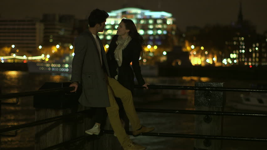 An attractive man and a woman are spending time together in London at night. They are overlooking the river Thames and the illuminated skyline. An illuminated barge passes through the shot.