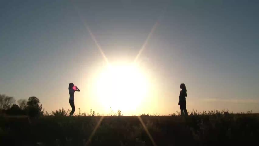 Two girls play catch on either side of large sun that is setting on horizon, atop a small hill with pink ball, and a blue sky horizon.