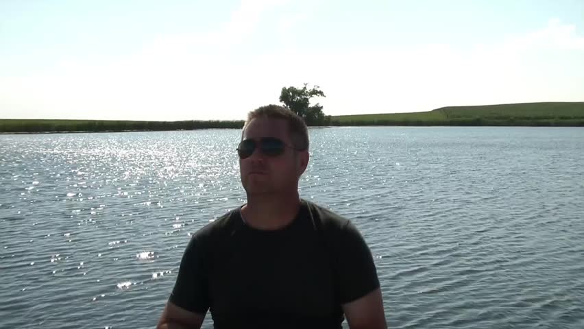 Man drinking a water bottle in summer sunshine with beautiful blue lake, and green fields in background.