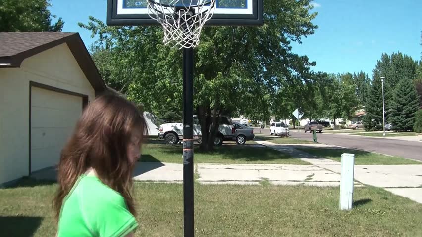 Young girl shoots baskets with her green basketball in driveway, outside in the sunshine.