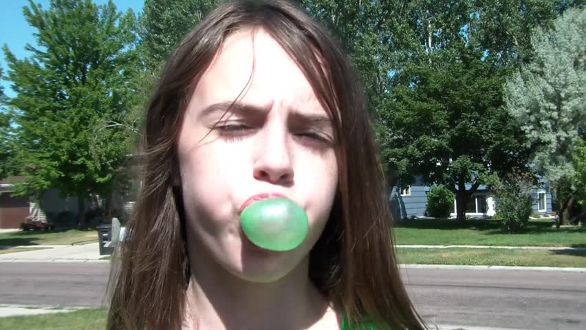 Girl blows bubble of gum at camera during sunny summer afternoon.