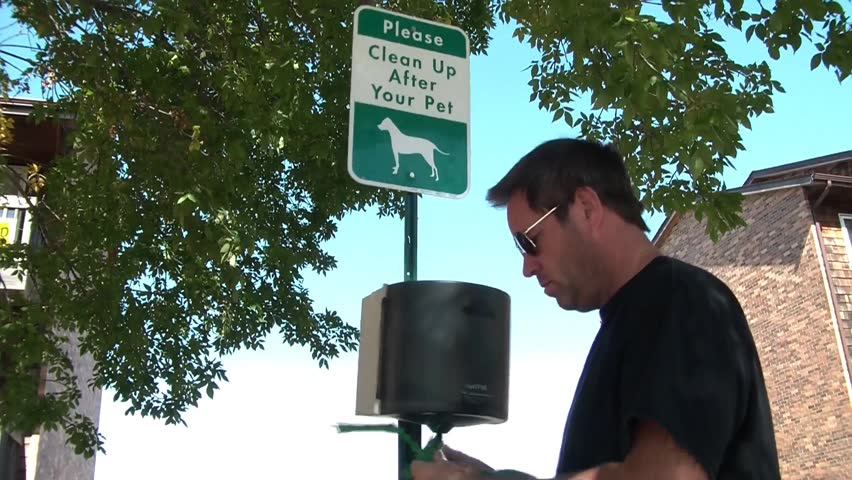 Clip of man pulling green bag from dog waste station reading please clean up after your pet, in middle of city during afternoon.