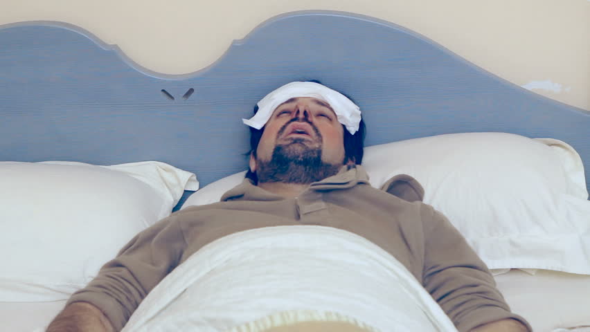 A man lies in the bed, he's got a bad flu / cold / illness. He looks at the thermometer, then he despairs because his body temperature is too high.