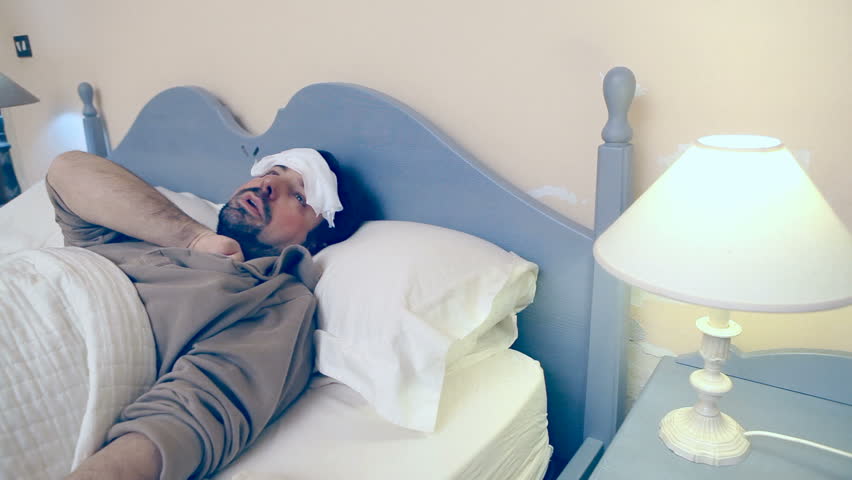 A man lies in the bed, he's got a bad flu / cold / illness. He looks at the thermometer, then he puts it near the heaten lamp to artificially raise the temperature.