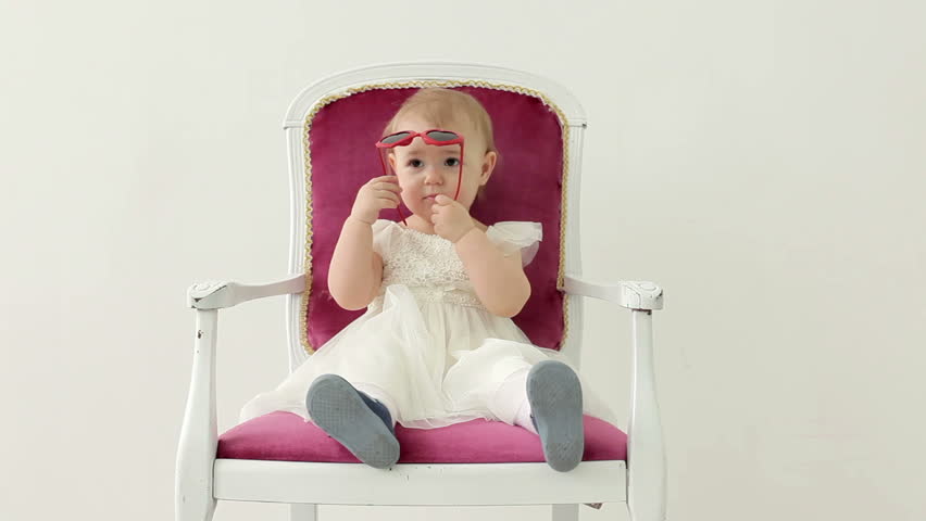 Little girl sitting in a vintage chair in studio and play with funny sunglasses. Timelaps