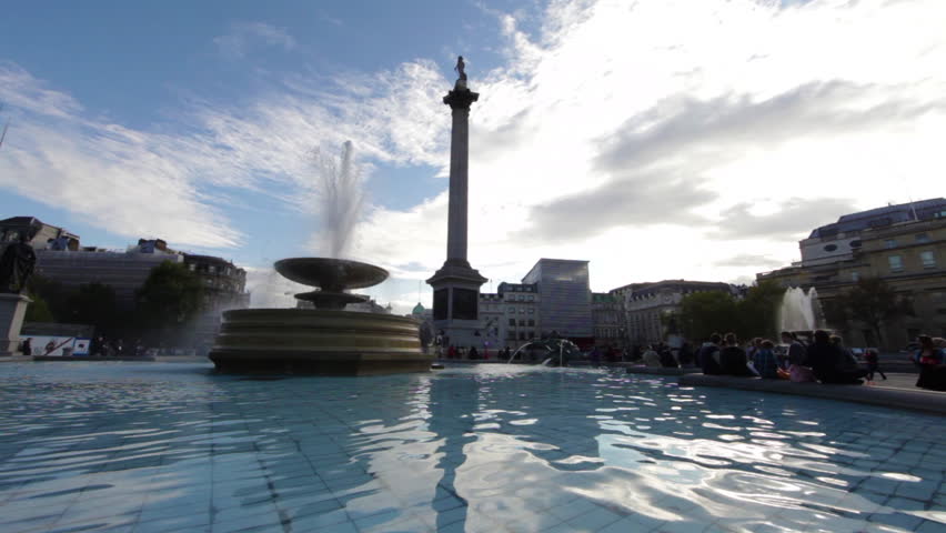 Stationary view of Lord Nelson's monument on Trafalgar Square in London. There is a fountain in the foreground. There are people sitting on the fountain. 