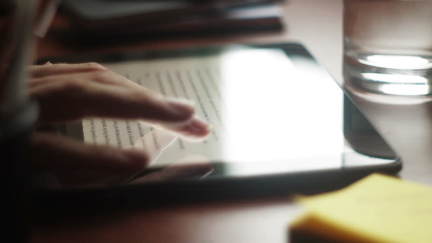 Hand flicks through a document of a tablet on a desk. Dolly shot to stationary, backlit.