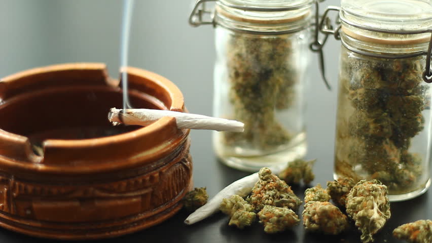 Marijuana Joint Burning 1. A marijuana filled joint burning on the edge of a wooden ashtray. Jars of marijuana and more around.